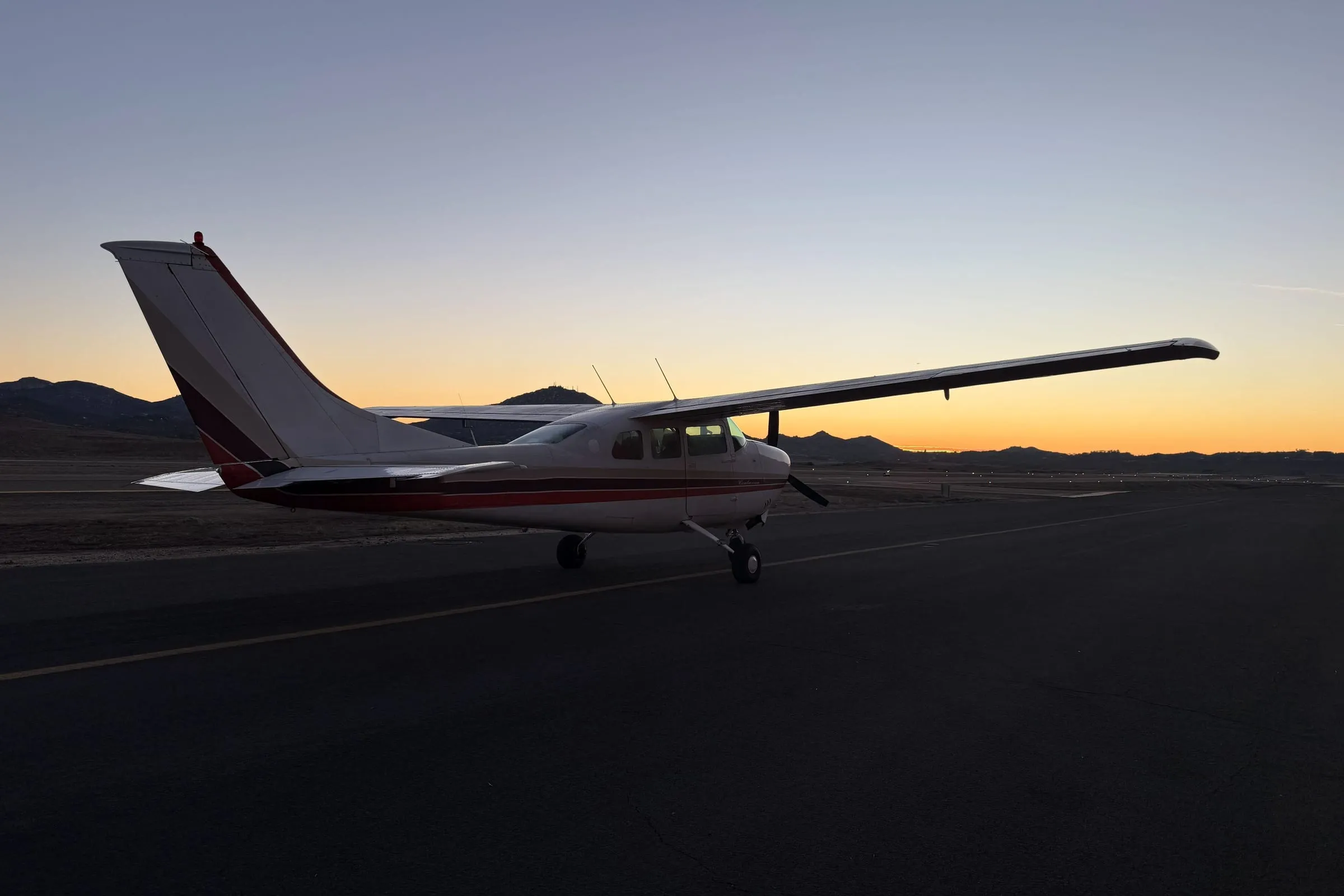 A high-wing single-engine airplane parked on a ramp at dusk, silhouetted against an orange sky and low hills