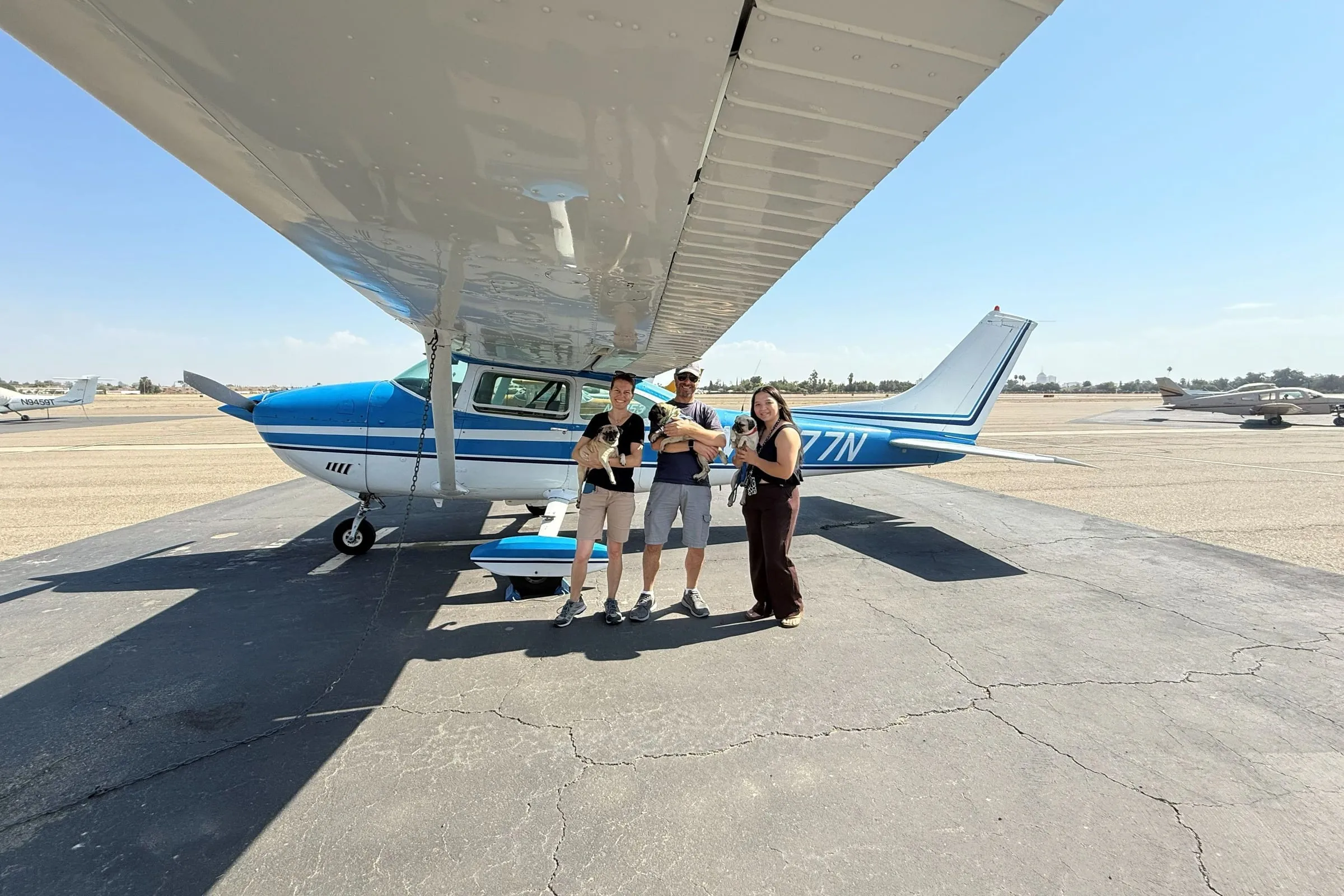 Three volunteers holding small rescue dogs under the wing of a blue-and-white Cessna on a sunny ramp