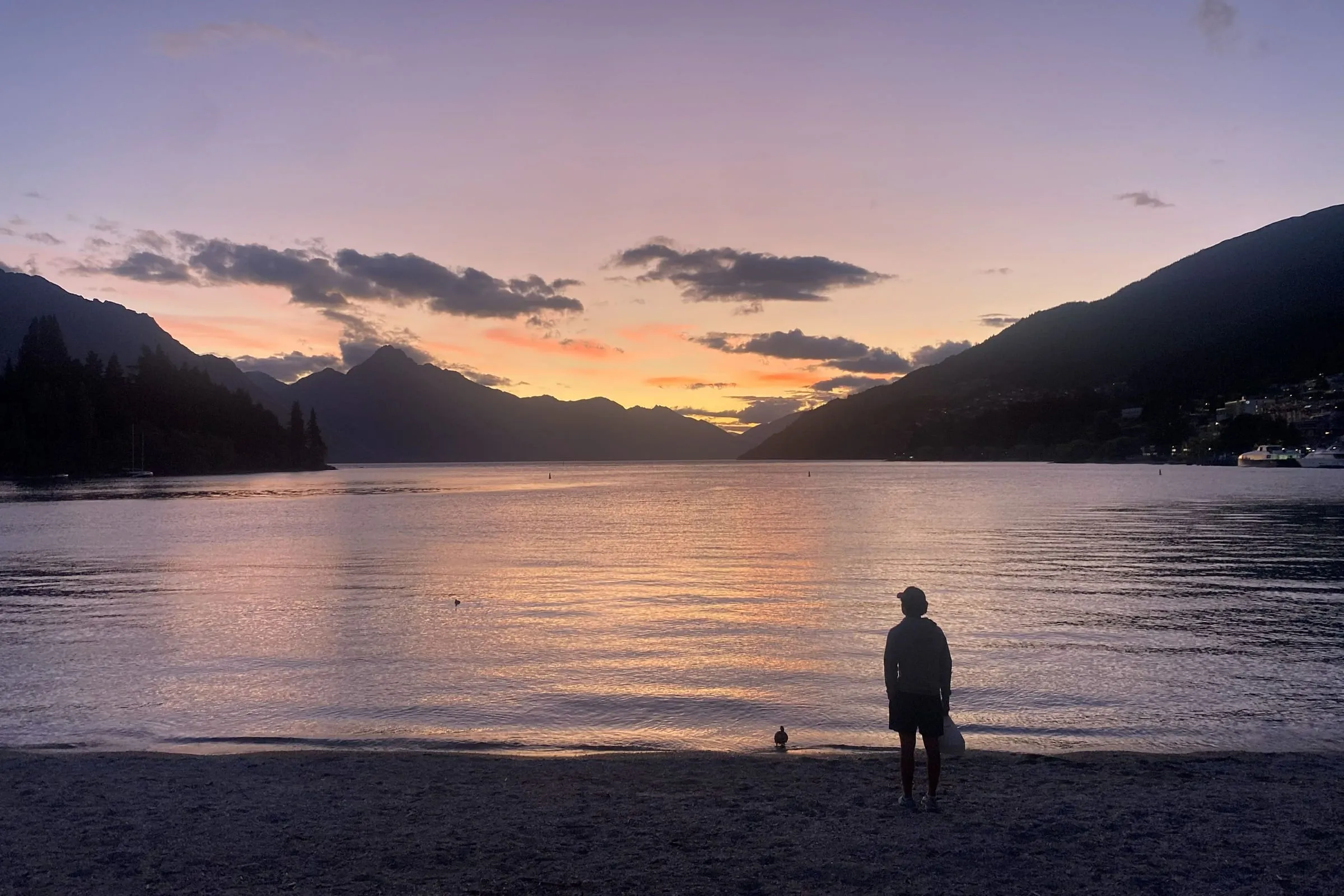 A lone figure on a pebble beach watching the sun go down over a mountain lake